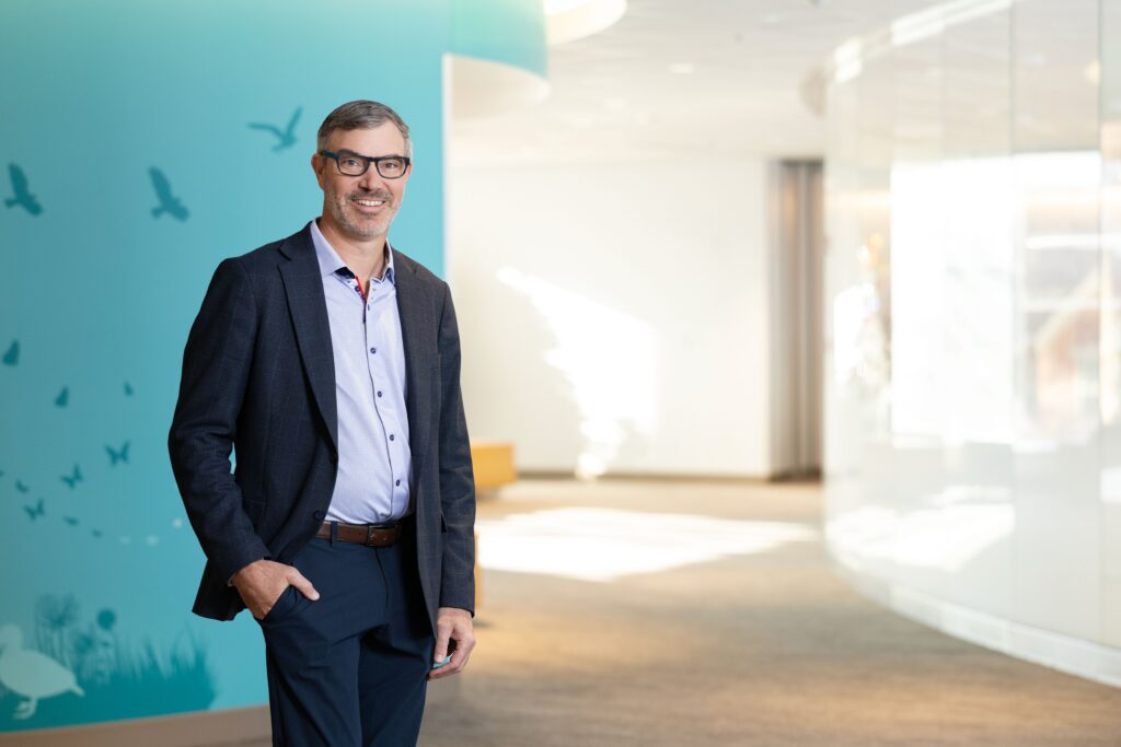 Ryan Goodwin, MD, MBA, standing in a hallway with a teal wall. He is wearing a blue shirt and black suit pants and jacket.