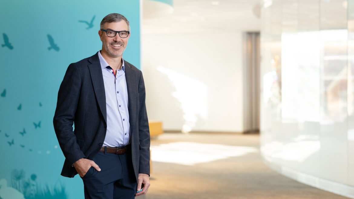 Ryan Goodwin, MD, MBA, standing in a hallway with a teal wall. He is wearing a blue shirt and black suit pants and jacket.