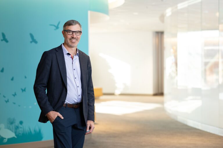 Ryan Goodwin, MD, MBA, standing in a hallway with a teal wall. He is wearing a blue shirt and black suit pants and jacket.