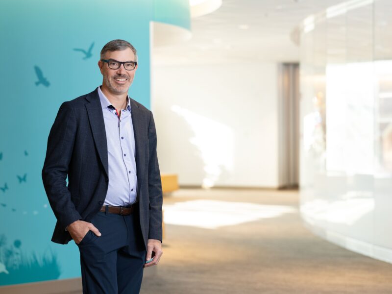 Ryan Goodwin, MD, MBA, standing in a hallway with a teal wall. He is wearing a blue shirt and black suit pants and jacket.