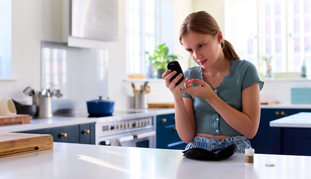 Young adult using a finger-prick device to check blood sugar levels while holding a glucose meter, standing at a kitchen counter with a small medical kit nearby.