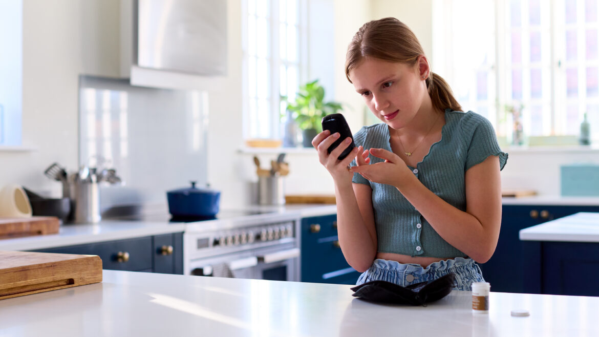 Young adult using a finger-prick device to check blood sugar levels while holding a glucose meter, standing at a kitchen counter with a small medical kit nearby.