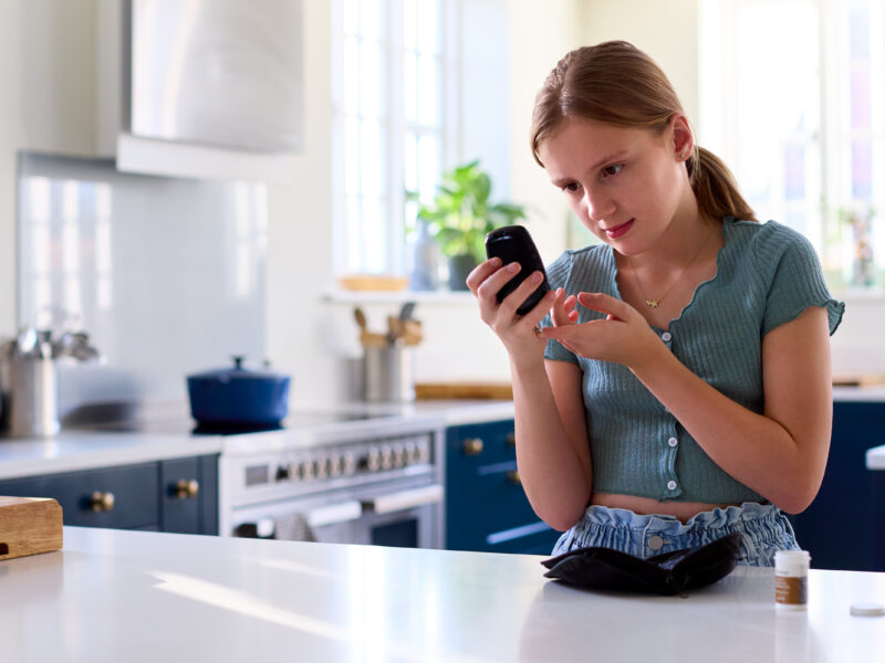 Young adult using a finger-prick device to check blood sugar levels while holding a glucose meter, standing at a kitchen counter with a small medical kit nearby.