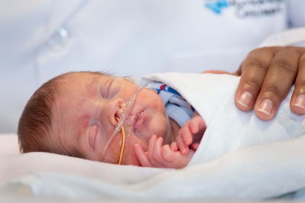 Baby in a NICU bed with nasal canula. Swaddled in white blanket with health care worker's hand gently resting on the baby.
