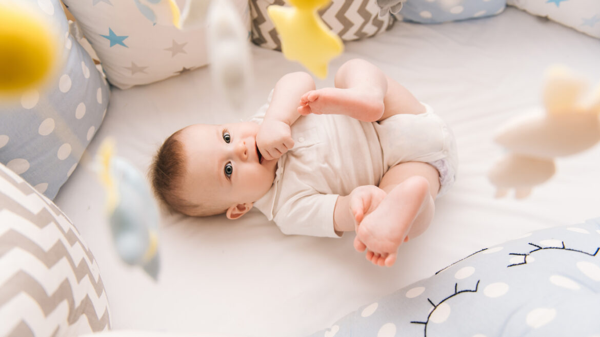 Cute baby lies in a white round bed. Light nursery for young children. Toys for infant cot. Smiling child playing with mobile of felt and chewing his feet in sunny bedroom.