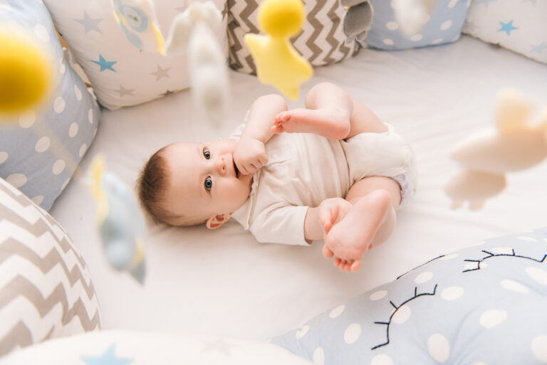 Cute baby lies in a white round bed. Light nursery for young children. Toys for infant cot. Smiling child playing with mobile of felt and chewing his feet in sunny bedroom.