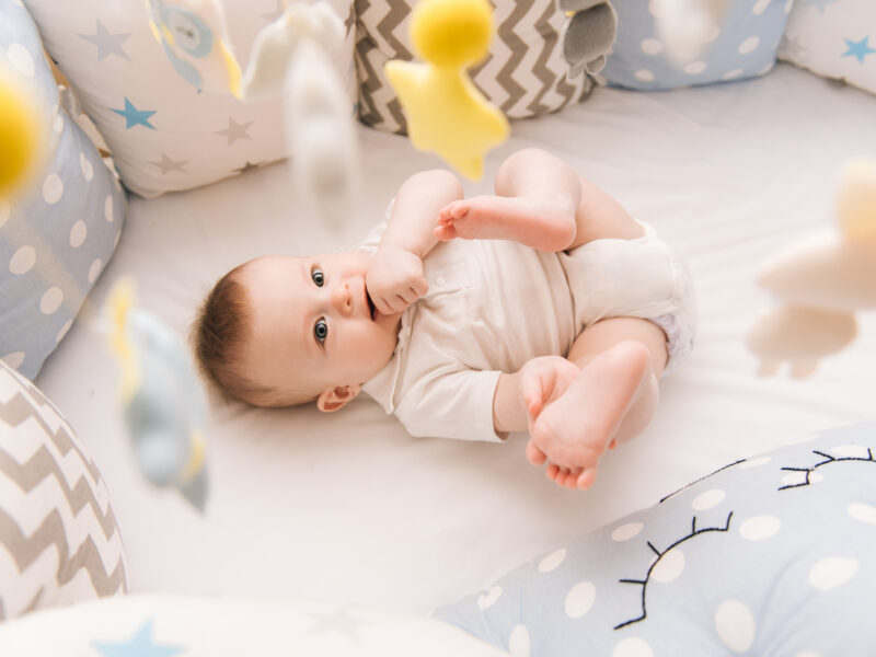 Cute baby lies in a white round bed. Light nursery for young children. Toys for infant cot. Smiling child playing with mobile of felt and chewing his feet in sunny bedroom.
