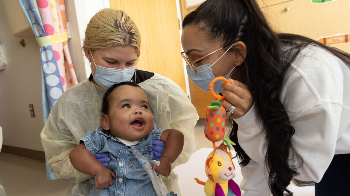 Baby with tracheostomy tube happily engaging with medical team who are holding a toy and wearing masks and gowns.