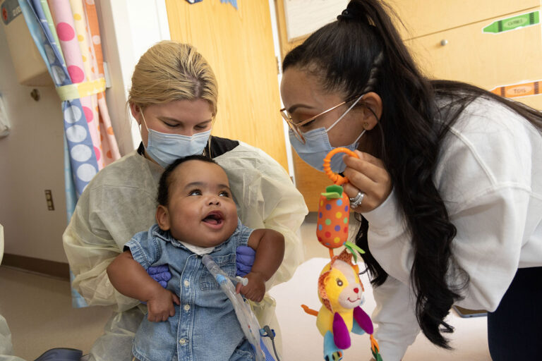 Baby with tracheostomy tube happily engaging with medical team who are holding a toy and wearing masks and gowns.