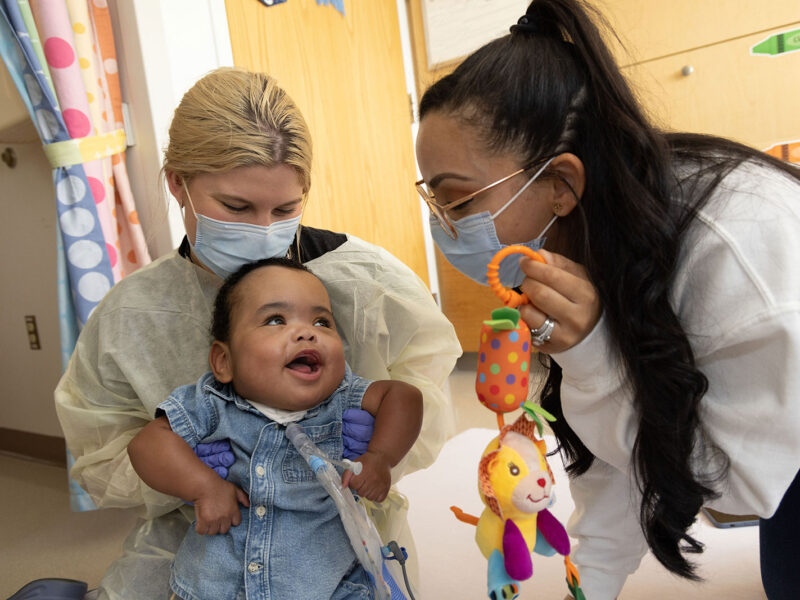 Baby with tracheostomy tube happily engaging with medical team who are holding a toy and wearing masks and gowns.