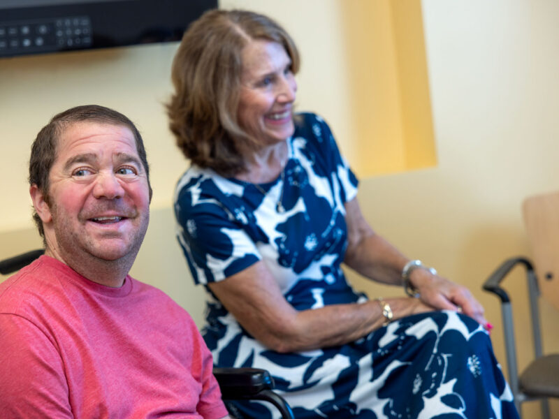 A smiling man with Duchenne muscular dystrophy sits beside a woman in a hospital room before or during treatment.