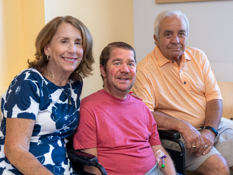 A man with Duchenne muscular dystrophy sits in a wheelchair between two family members while waiting for an intravenous infusion.