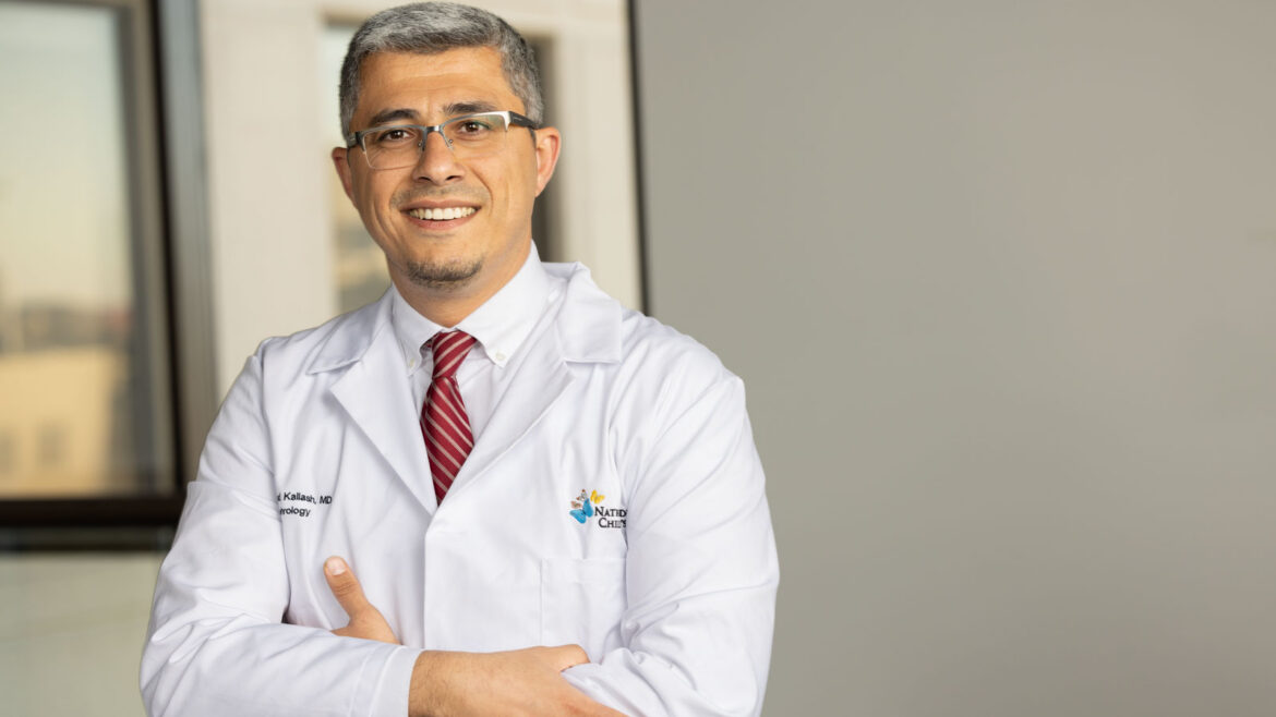 Dr. Kallash standing in an office hallway, wearing a white coat and red tie.