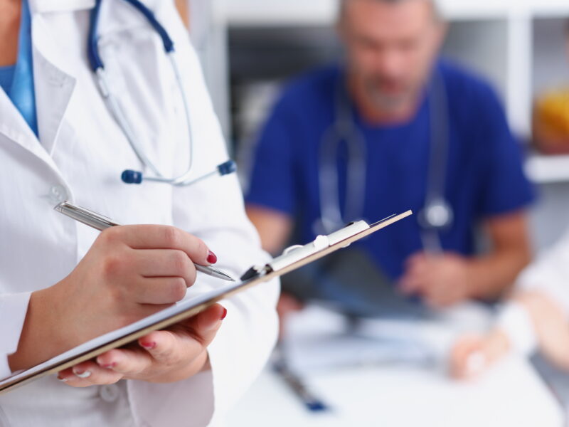 Doctor in a white coat holding a clipboard pad and fill medical history list with silver pen.