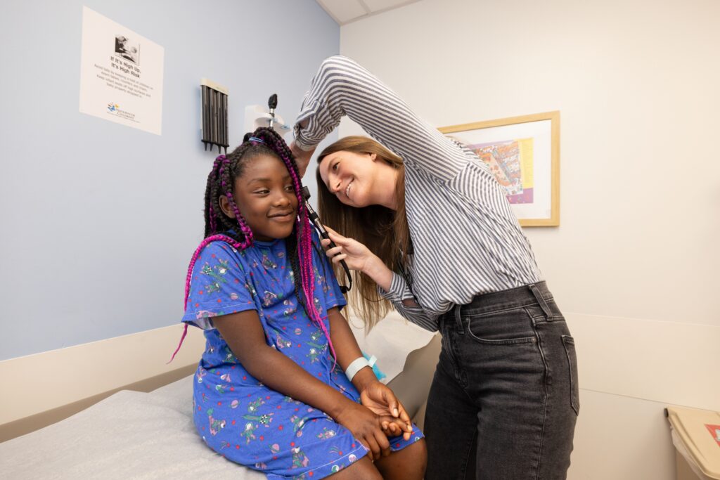 A smiling girl sits on a table at a doctor's office. A smiling female doctor is checking the girl's ear.