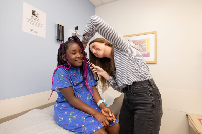 A smiling girl sits on a table at a doctor's office. A smiling female doctor is checking the girl's ear.