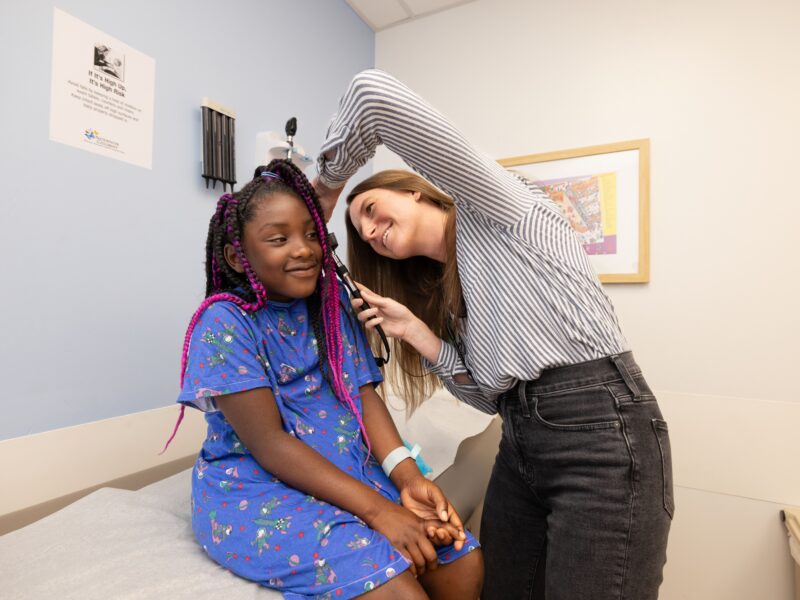 A smiling girl sits on a table at a doctor's office. A smiling female doctor is checking the girl's ear.