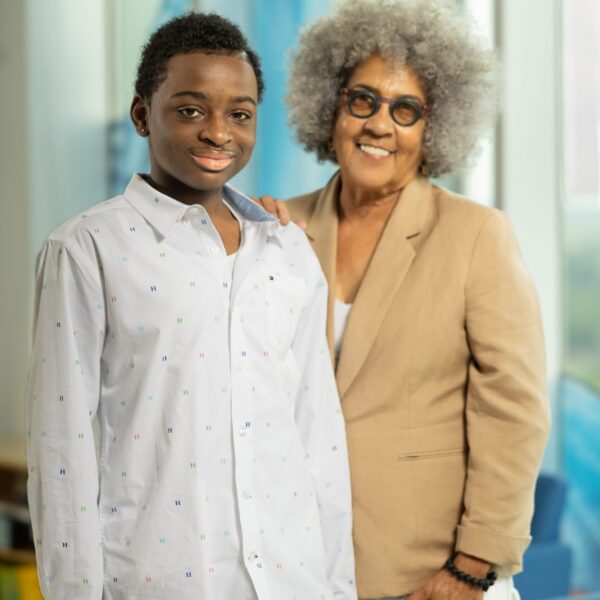 A teenager in a white shirt stand next to a woman wearing a tan jacket. They are both looking toward the camera.