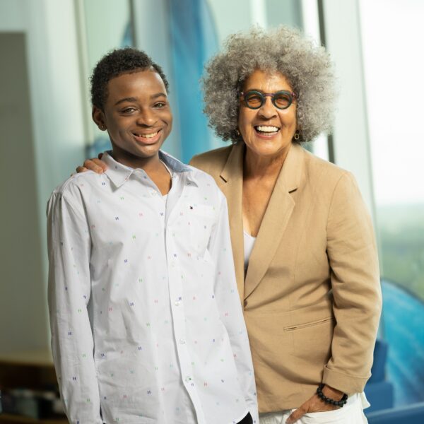 A teenager in a white shirt stand next to a woman wearing a tan jacket. They are both looking toward the camera.