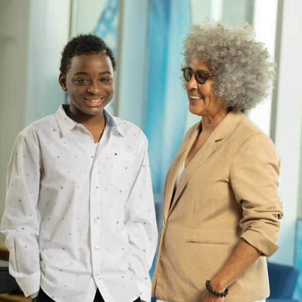 A teenager in a white shirt stand next to a woman wearing a tan jacket who is looking at him while he looks at the camera.