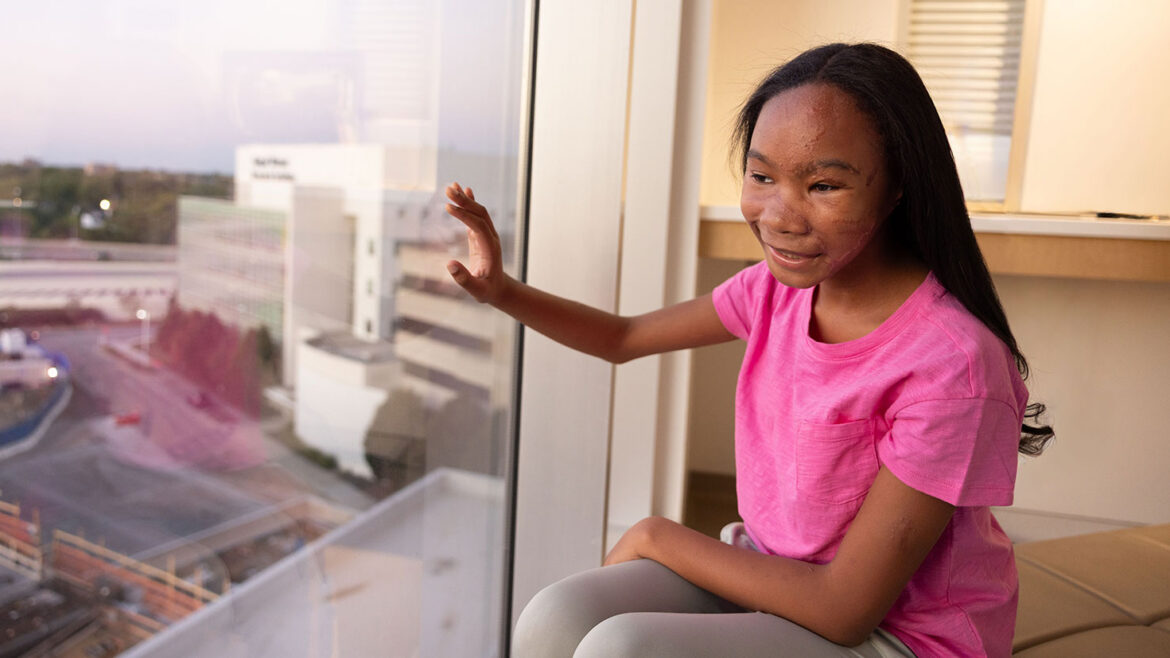 A girl in a bright pink shirt smiles while sitting beside a large hospital window and looking outside.