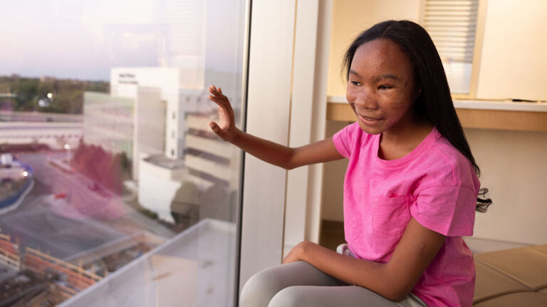 A girl in a bright pink shirt smiles while sitting beside a large hospital window and looking outside.
