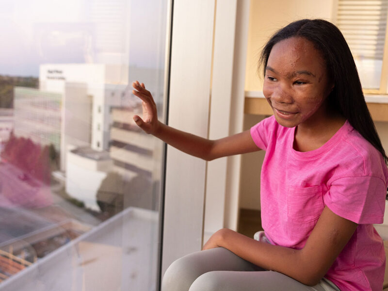 A girl in a bright pink shirt smiles while sitting beside a large hospital window and looking outside.